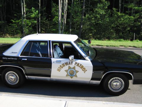 1991 Ford LTD Crown Victoria - Close profile of a black-and-white Ford police sedan with HIGHWAY PATROL lettering, CHP star, and red door-mounted spotlight.