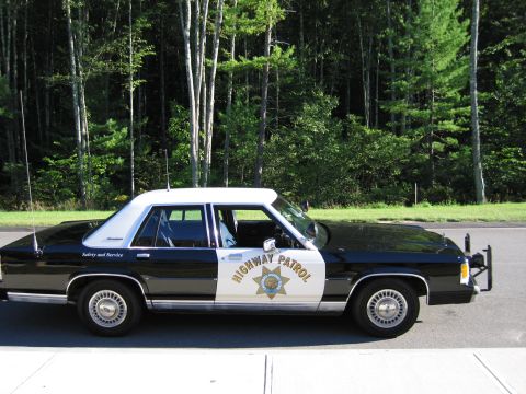 1991 Ford LTD Crown Victoria - Profile of a black-and-white Ford police sedan marked HIGHWAY PATROL with gold star emblem, parked on pavement beside trees.