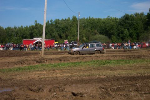 1979 Ford F-150 - A silver SUV drives through a muddy course during an event, with a crowd watching from behind a barrier.