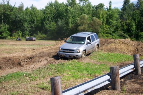 1979 Ford F-150 - An SUV is stuck in a muddy trail, with trees and fields surrounding it. Another car and an ATV are in the distance.