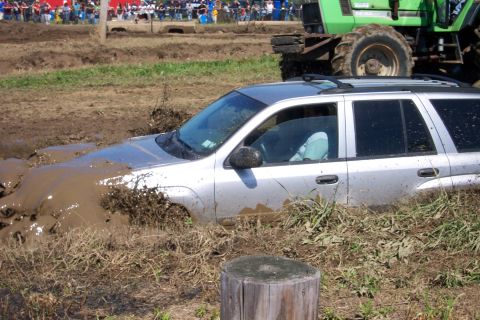 1979 Ford F-150 - A silver SUV is deeply stuck in mud, with a large green tractor visible behind it in a muddy field.