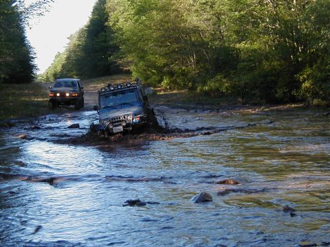 1993 Ford Bronco - A blue Jeep Wrangler with a roof-mounted light bar and tubular front bumper wades nose-down through a deep, muddy water crossing on a woode…