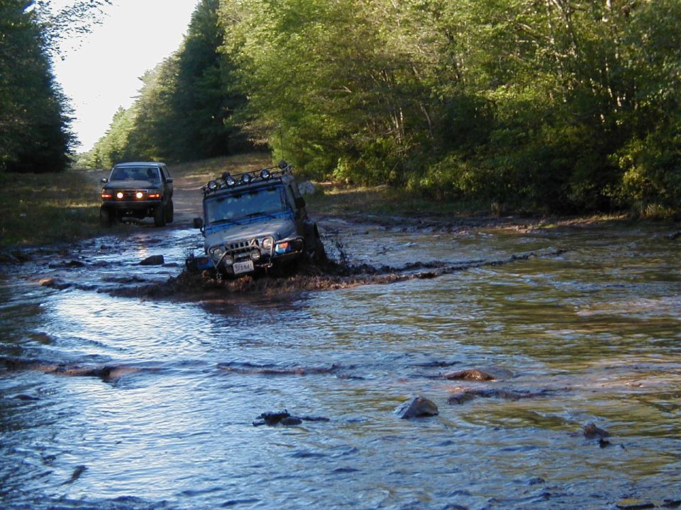 Blue Jeep Wrangler with roof light bar crossing deep muddy water on a forest trail