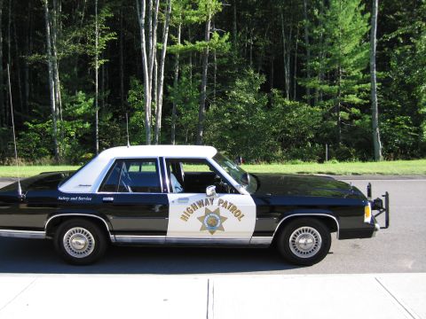 1991 Ford LTD Crown Victoria - Profile of a black-and-white Ford police sedan with HIGHWAY PATROL lettering and CHP star emblem, parked near a pine forest.