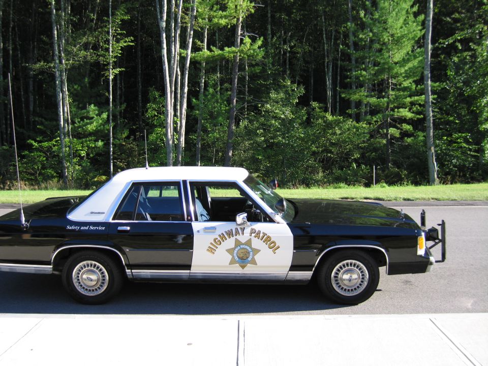 Side view of black-and-white Ford Highway Patrol sedan by woods