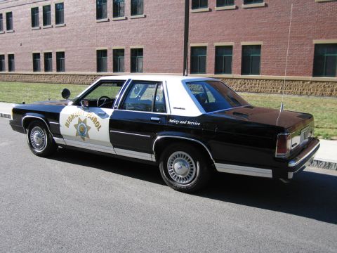 1991 Ford LTD Crown Victoria - Rear three-quarter view of a black-and-white Ford police sedan marked HIGHWAY PATROL and Safety and Service, parked by a brick building.
