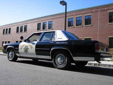 1991 Ford LTD Crown Victoria - Rear three-quarter view of a black-and-white Ford police sedan with Safety and Service lettering, parked beside a brick building.