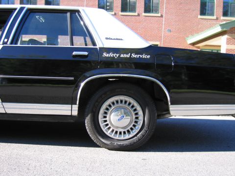1991 Ford LTD Crown Victoria - Close-up of a black sedan's rear door and Ford-hubcap wheel, with white Safety and Service lettering, by a brick building.
