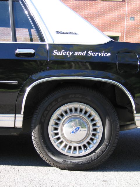 1991 Ford LTD Crown Victoria - Close-up of a black sedan's rear wheel with a Ford-logo hubcap and Goodyear tire, below white Safety and Service lettering.