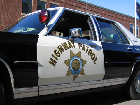 1991 Ford LTD Crown Victoria - Angled view of a black-and-white police sedan's front door with HIGHWAY PATROL lettering and CHP star, parked by a brick building.