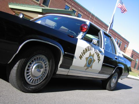 1991 Ford LTD Crown Victoria - Low-angle side view of a black-and-white Ford police sedan marked HIGHWAY PATROL, an American flag flying behind a brick building.