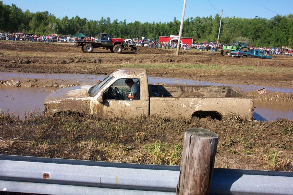 White pickup truck stuck deep in mud, driver visible inside. Other vehicles and spectators in background at a mud bog event