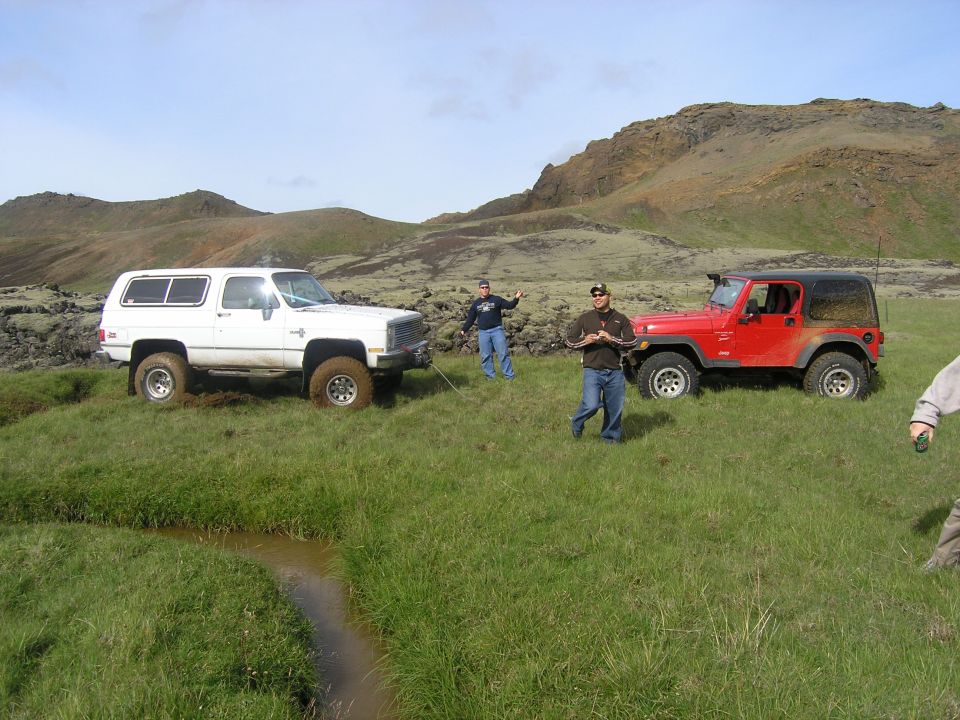 White Chevrolet K5 Blazer and red Jeep Wrangler parked in a field with people nearby