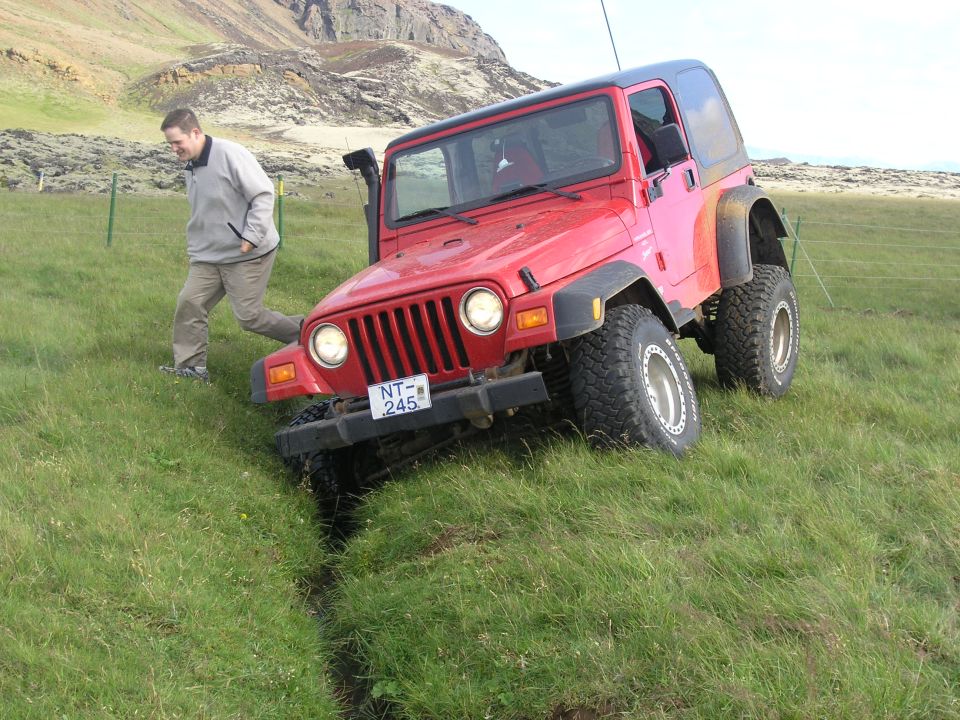 Red Jeep TJ driving off-road on a grassy hill with a man walking nearby. License plate NT-245