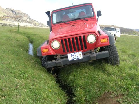 2001 Jeep Wrangler - A red Jeep Wrangler navigates a narrow ditch in a grassy field, with another vehicle visible in the background.