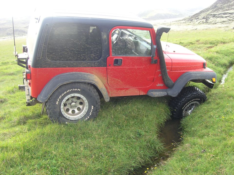Red 2001 Jeep TJ with snorkel stuck in a grassy ditch, front wheel submerged