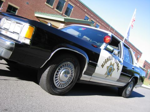 1991 Ford LTD Crown Victoria - Low-angle front three-quarter view of a black-and-white Ford police sedan marked HIGHWAY PATROL, with a flag and brick building behind.