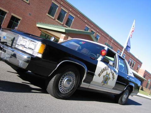 1991 Ford LTD Crown Victoria - Low-angle front three-quarter view of a black-and-white Ford police sedan marked HIGHWAY PATROL, parked near a flagpole and brick building.