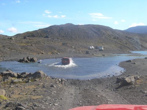 2001 Jeep Wrangler - A red SUV navigates a water crossing in a rocky, barren landscape under a clear blue sky.