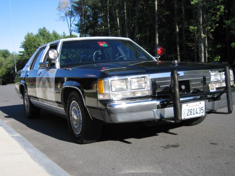1991 Ford LTD Crown Victoria - Front three-quarter shot of a black Ford police-replica sedan with chrome trim, push bumper, and rooftop beacon beside a forest road.