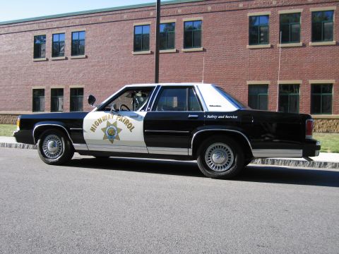 1991 Ford LTD Crown Victoria - Side view of a black-and-white Highway Patrol replica sedan parked along the curb in front of a brick building.