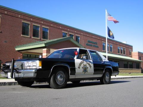1991 Ford LTD Crown Victoria - black-and-white Highway Patrol replica sedan parked in front of a brick middle school with U.S. and state flags flying.