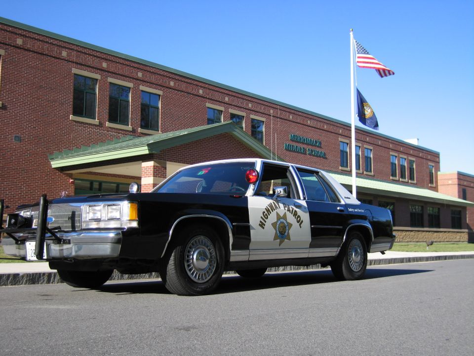Black-and-white Highway Patrol sedan parked outside brick school with flag