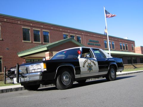 1991 Ford LTD Crown Victoria - Front-quarter view of a black-and-white Highway Patrol replica sedan with a push bumper parked outside a flag-flying brick school.