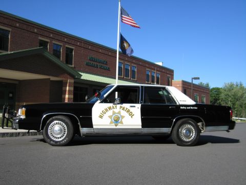 1991 Ford LTD Crown Victoria - Side profile of a black-and-white Highway Patrol replica sedan parked in front of a brick school with flags overhead.