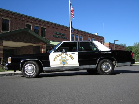 1991 Ford LTD Crown Victoria - Front three-quarter view of a black-and-white Highway Patrol replica sedan parked at a brick school with flags flying.
