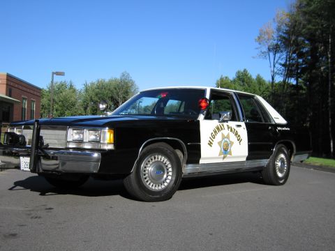 1991 Ford LTD Crown Victoria - Front three-quarter view of a black-and-white Ford Highway Patrol replica sedan with a push bumper on an open lot by trees.