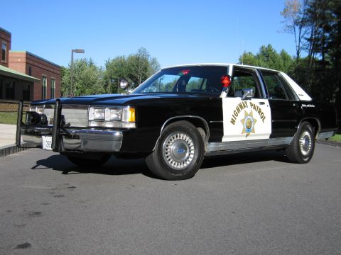 1991 Ford LTD Crown Victoria - Front three-quarter view of a black-and-white Ford Highway Patrol replica sedan with a push bumper and roof beacon.