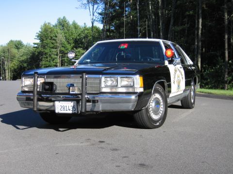 1991 Ford LTD Crown Victoria - Front view of a black-and-white Ford police-replica sedan with a chrome grille, brush-guard bumper, and red roof beacon.
