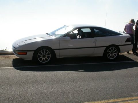 1990 Ford Probe - A white 1990 Ford Probe is parked on the side of a road with two people standing behind it.