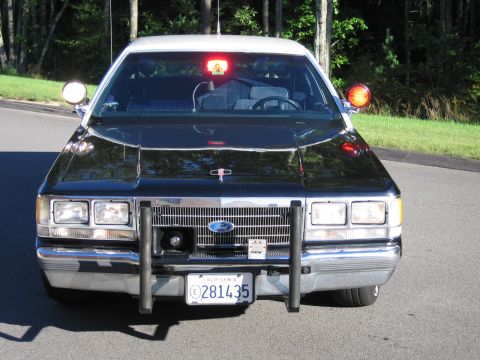 1991 Ford LTD Crown Victoria - Head-on front view of a black Ford police-replica sedan with door spotlights, a red roof beacon, and a push bumper.
