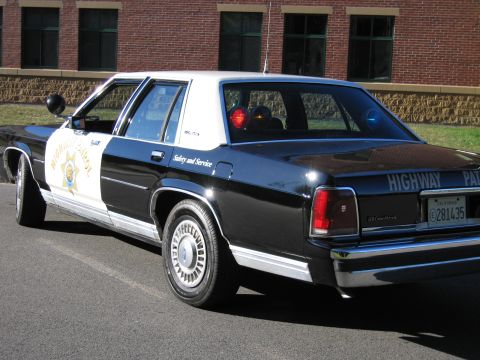 1991 Ford LTD Crown Victoria - Rear three-quarter view of a black-and-white Ford Highway Patrol replica sedan with trunk lettering, parked by a brick wall.