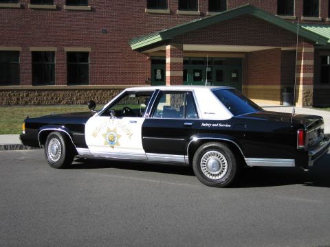 1991 Ford LTD Crown Victoria - Side rear-quarter view of a black-and-white Highway Patrol replica sedan parked outside a brick school building.