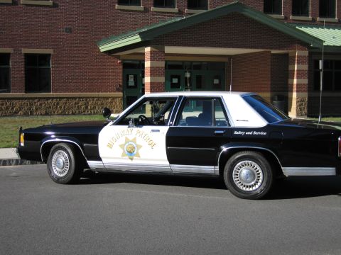 1991 Ford LTD Crown Victoria - Side profile of a black-and-white Highway Patrol replica sedan with door star emblem parked outside a brick school.