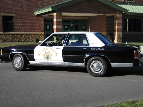 1991 Ford LTD Crown Victoria - Side profile of a black-and-white Highway Patrol replica sedan with HIGHWAY PATROL door lettering parked by a school entrance.