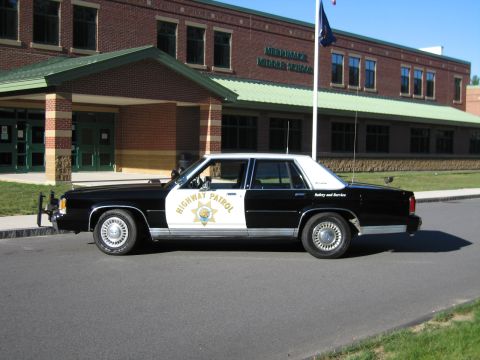 1991 Ford LTD Crown Victoria - black-and-white police sedan marked Highway Patrol Safety and Service sits in side profile outside a brick middle school building.