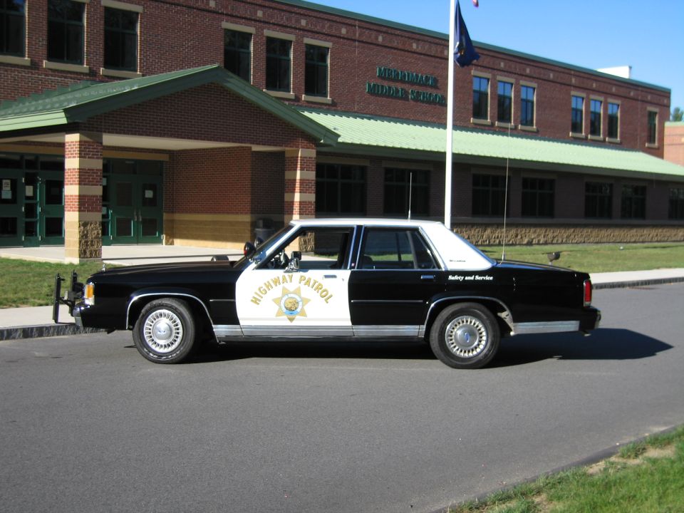 Black-and-white Highway Patrol police sedan parked in profile in front of a brick middle school