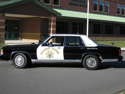 1991 Ford LTD Crown Victoria - Driver-side profile of a black-and-white Highway Patrol police sedan with a gold seven-point star door emblem, parked near a brick school.