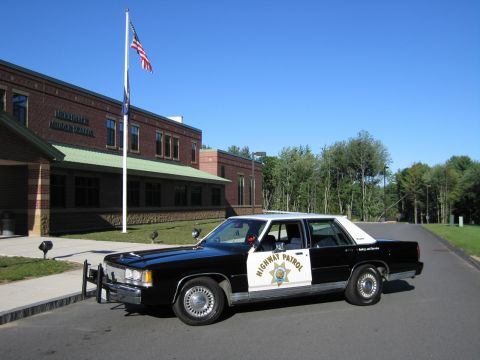 1991 Ford LTD Crown Victoria - black-and-white Highway Patrol police sedan parks beside a flagpole flying the US flag in front of a brick middle school.