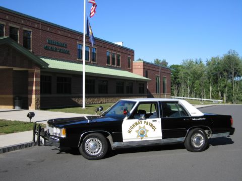 1991 Ford LTD Crown Victoria - Front three-quarter view of a black-and-white Highway Patrol police sedan with a push bar, parked outside a brick school under a clear sky.