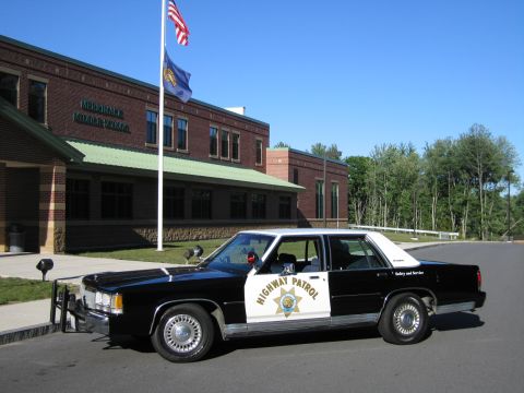 1991 Ford LTD Crown Victoria - black-and-white Highway Patrol police sedan with a front push bar sits outside a brick middle school beside a flagpole.