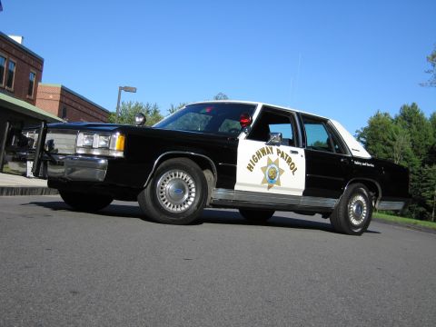 1991 Ford LTD Crown Victoria - Low-angle front three-quarter shot of a black-and-white Highway Patrol police sedan with a Ford wheel center, on a paved lot.