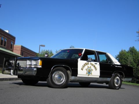 1991 Ford LTD Crown Victoria - Low front-quarter view of a black-and-white Highway Patrol police sedan showing red and amber roof beacons against a blue sky.
