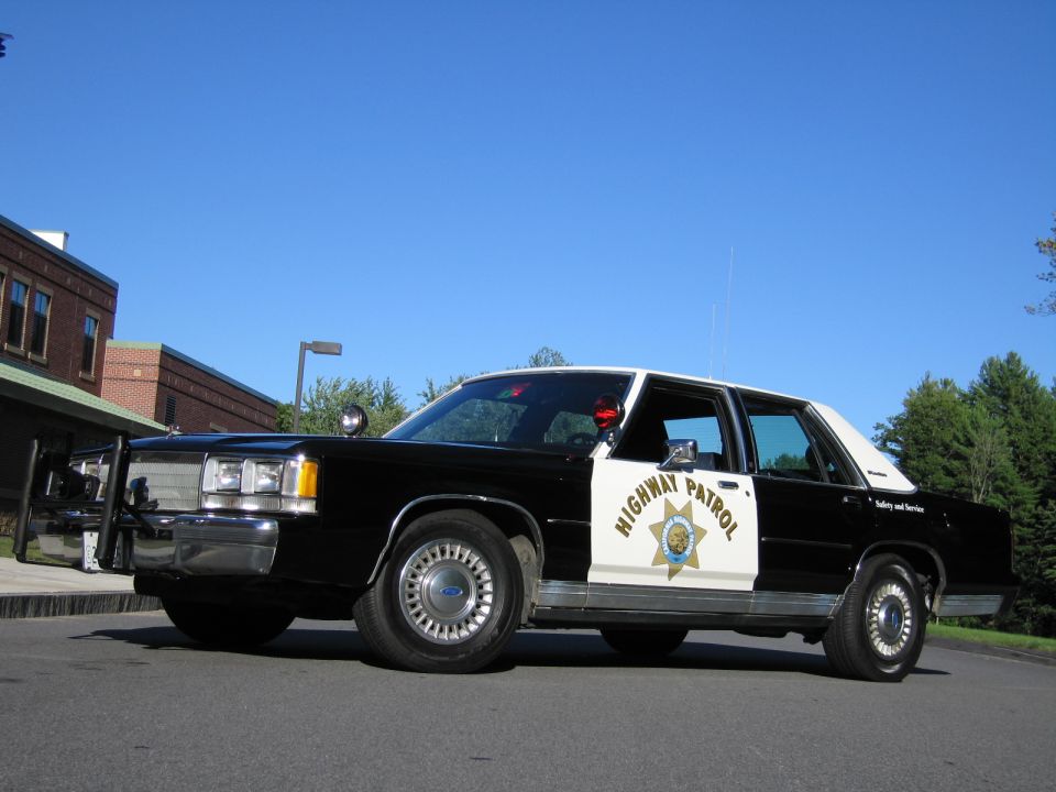 Low front-quarter view of a black-and-white Highway Patrol police sedan with roof lights