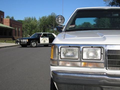 1991 Ford LTD Crown Victoria - silver car's front end and headlights dominate the frame as a black-and-white Highway Patrol sedan parks in the background.