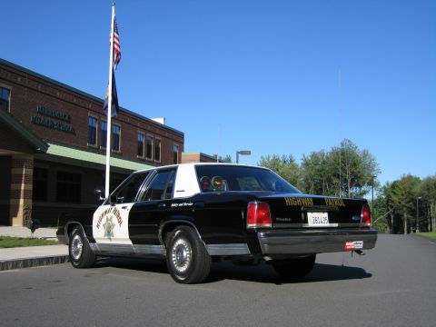 1991 Ford LTD Crown Victoria - Rear three-quarter view of a black-and-white Highway Patrol police sedan with a California plate, parked outside a brick school near a flag…